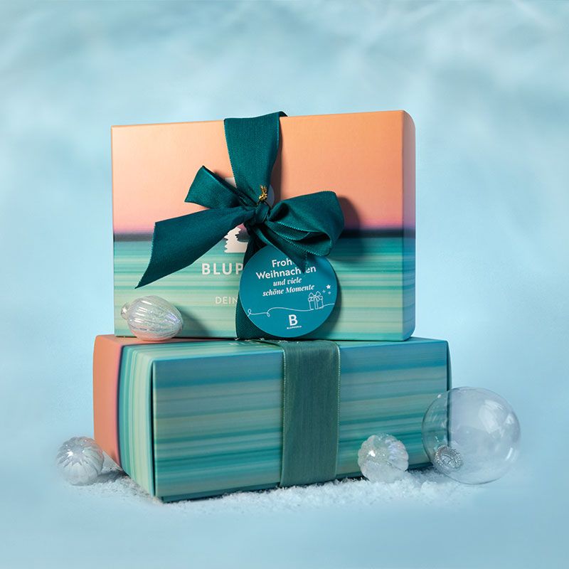 Bluphoria Geschenkboxen Zwei bunte Boxen mit Schleifen liegen aufeinander, daneben liegen Kunstschnee und Weihnachtskugeln.