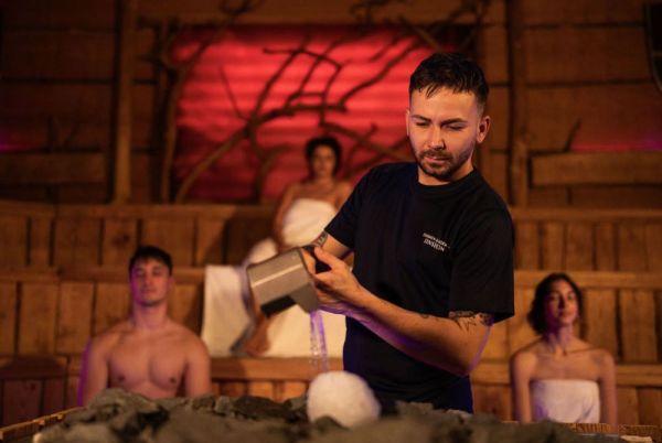 A man pours water into a sauna with three people in the background